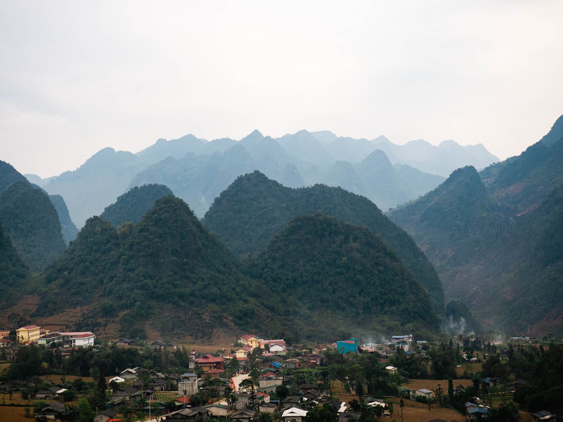 Green peaks rising behind a village in a valley, layers of mountains fading into haze