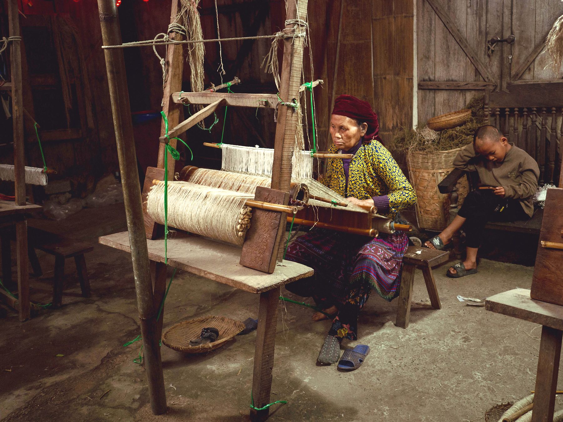 An elderly woman weaving at a wooden loom inside a dim traditional house