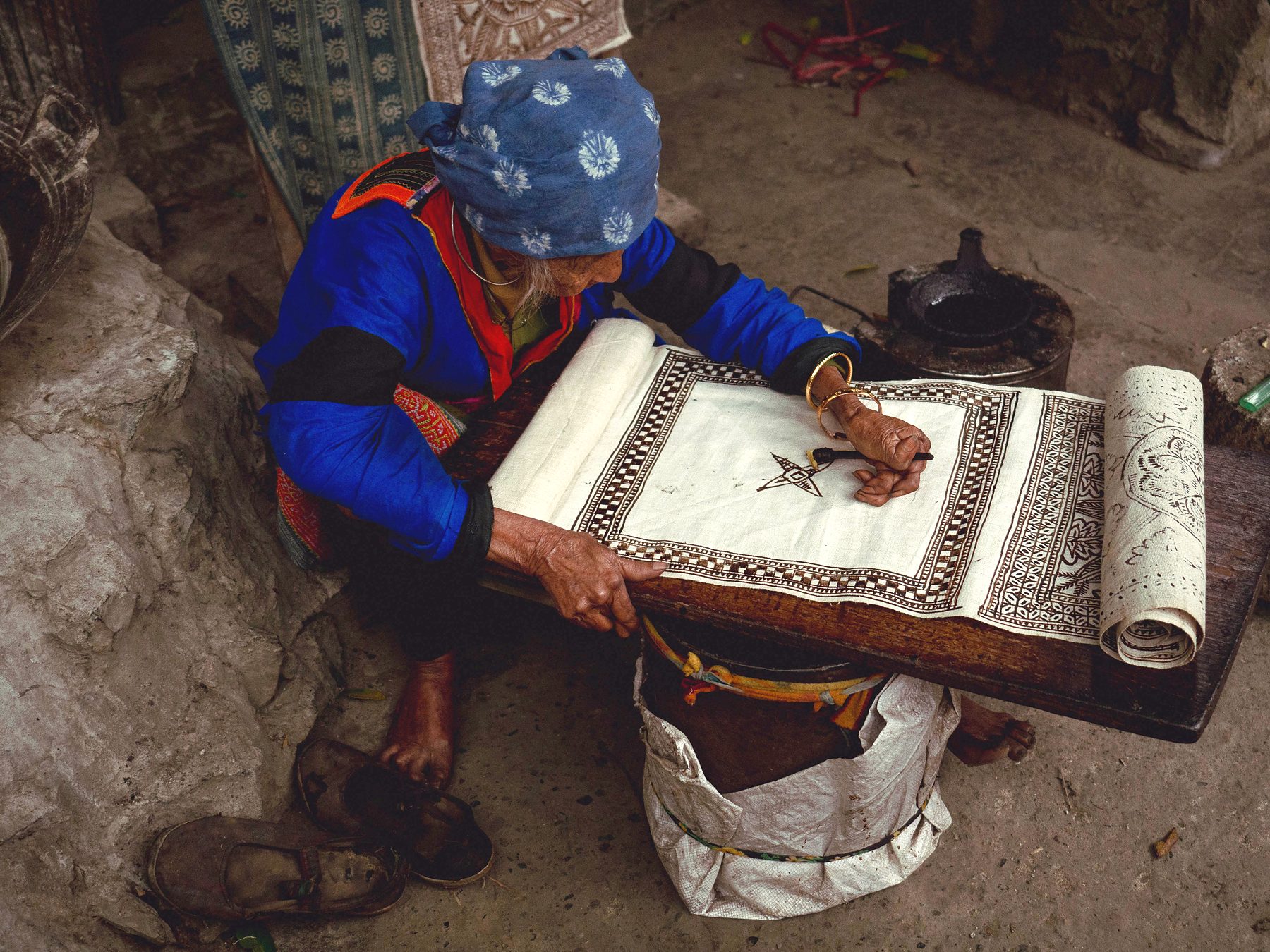 An elderly woman in a blue headscarf hand-embroidering a long patterned cloth