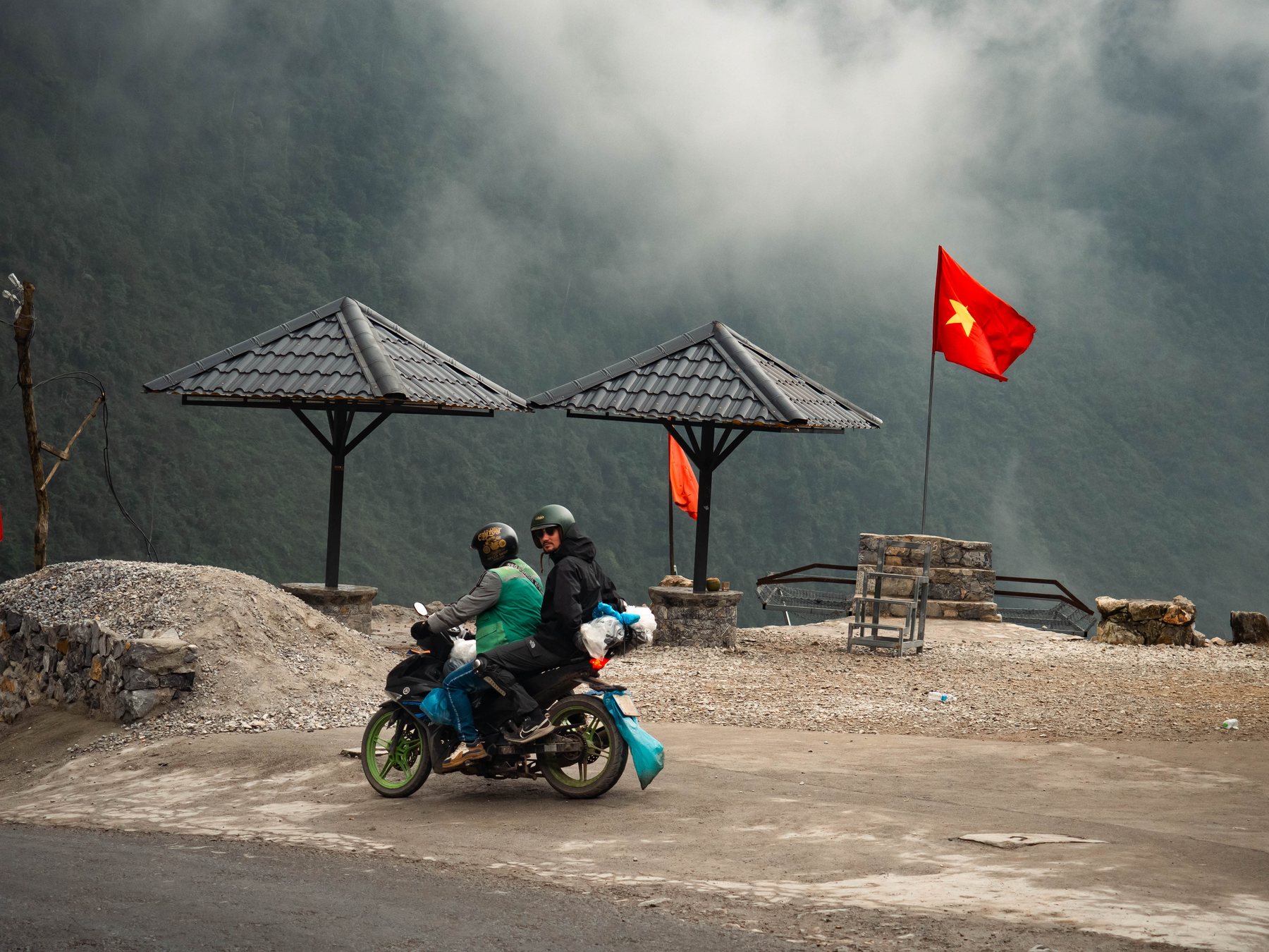A motorbike with two riders at a roadside viewpoint with a Vietnamese flag under heavy cloud